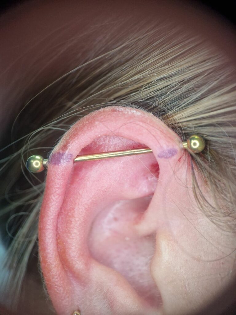 Piercing industriel avec barre dorée traversant le haut de l’oreille, réalisé par Oc Piercing au salon Jolie Piqûre à Arles