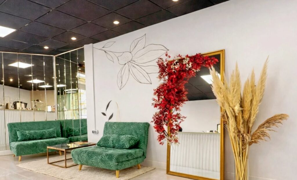 Espace d’attente du salon Jolie Piqûre à Arles : canapés en velours vert, grand miroir doré, fleurs rouges et pampas