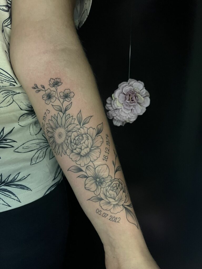 Tatouage fineline sur l’avant-bras : bouquet de tournesol, pivoines, orchidée et fleurs de cerisier accompagné de dates, réalisé à Arles
