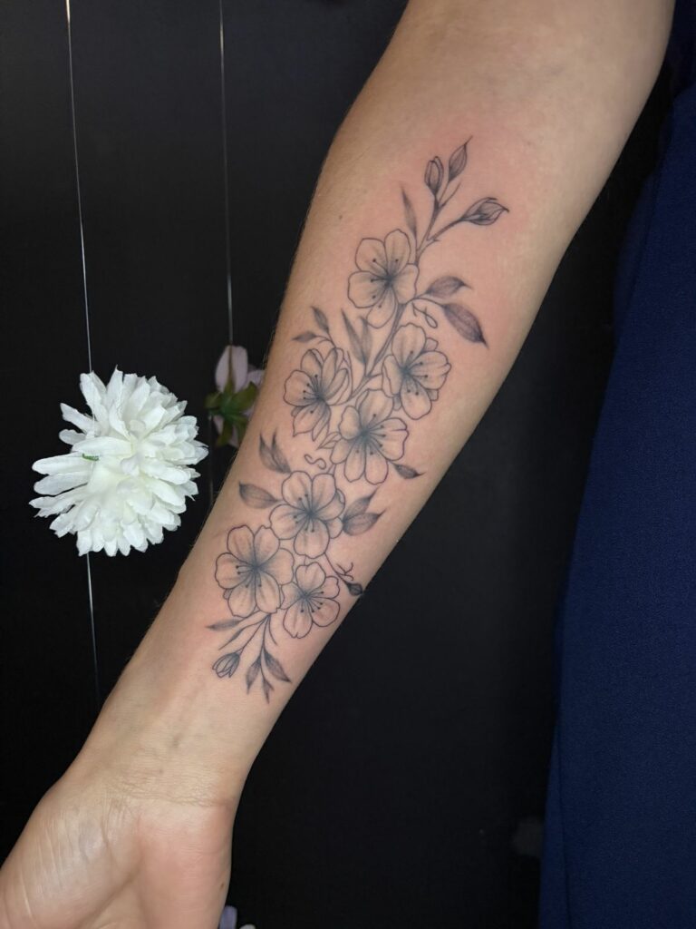 Tatouage fineline sur l’avant-bras : branche de fleurs de cerisier (sakura) aux pétales ombrés, boutons et feuilles, réalisé au salon Jolie Piqûre à Arles