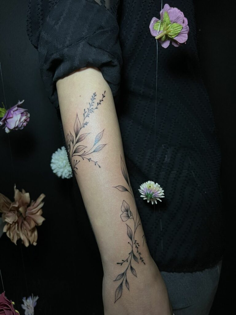 Tatouage fineline de branches fleuries et feuillages qui s’enroulent autour de l’avant-bras, réalisé au salon Jolie Piqûre à Arles