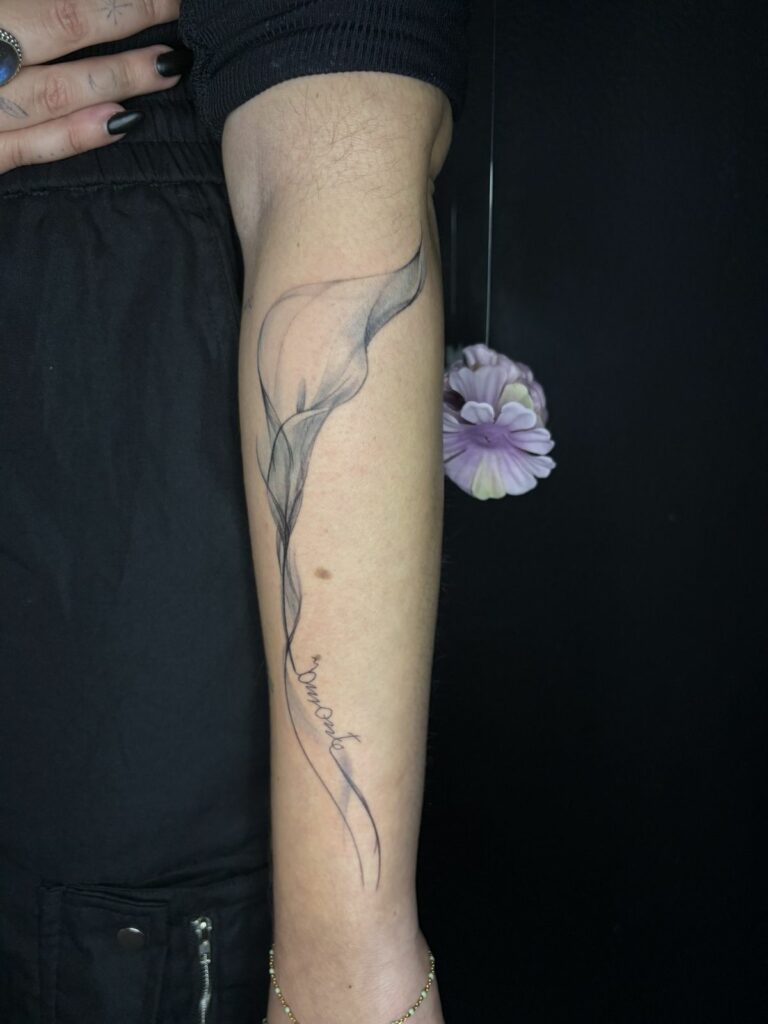 Tatouage fineline sur l’avant-bras : fleur d’arum aux ombrages légers façon aquarelle prolongée par une écriture manuscrite, réalisé au salon Jolie Piqûre à Arles
