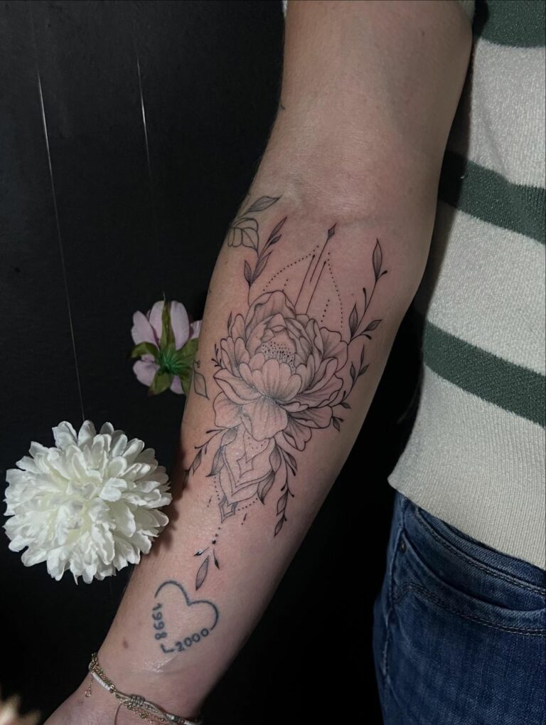 Tatouage fineline sur l’avant-bras : pivoine entourée de feuillages et d’ornements en pointillés, réalisé au salon Jolie Piqûre à Arles