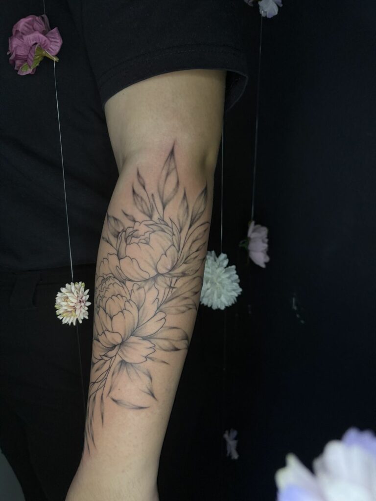 Tatouage fineline sur l’avant-bras : deux pivoines et leur feuillage aux ombrages délicats, réalisé au salon Jolie Piqûre à Arles