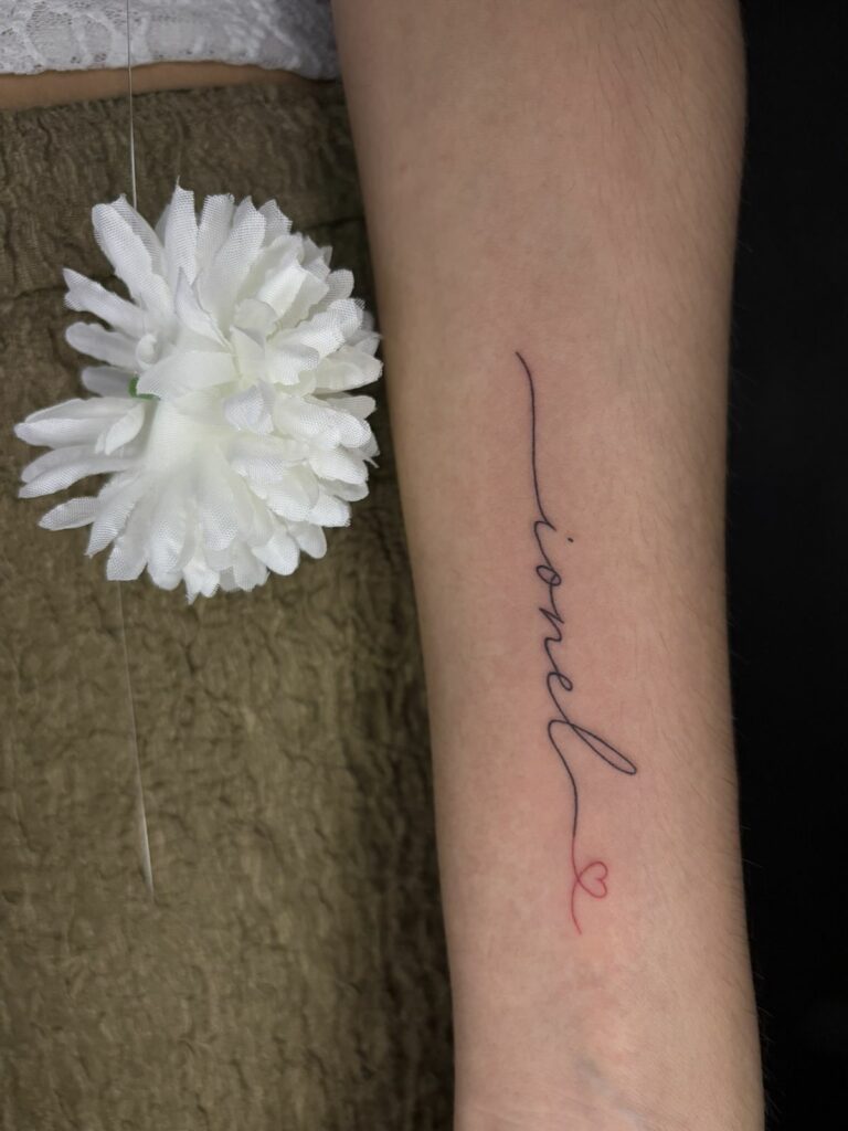 Tatouage lettering fineline vertical sur l’avant-bras : prénom en écriture manuscrite terminé par un petit cœur à l’encre rouge, réalisé au salon Jolie Piqûre à Arles