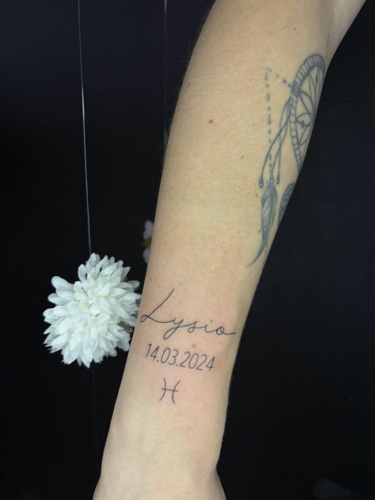 Tatouage fineline sur l’avant-bras : prénom en écriture manuscrite, date de naissance et symbole astrologique Poissons, réalisé au salon Jolie Piqûre à Arles