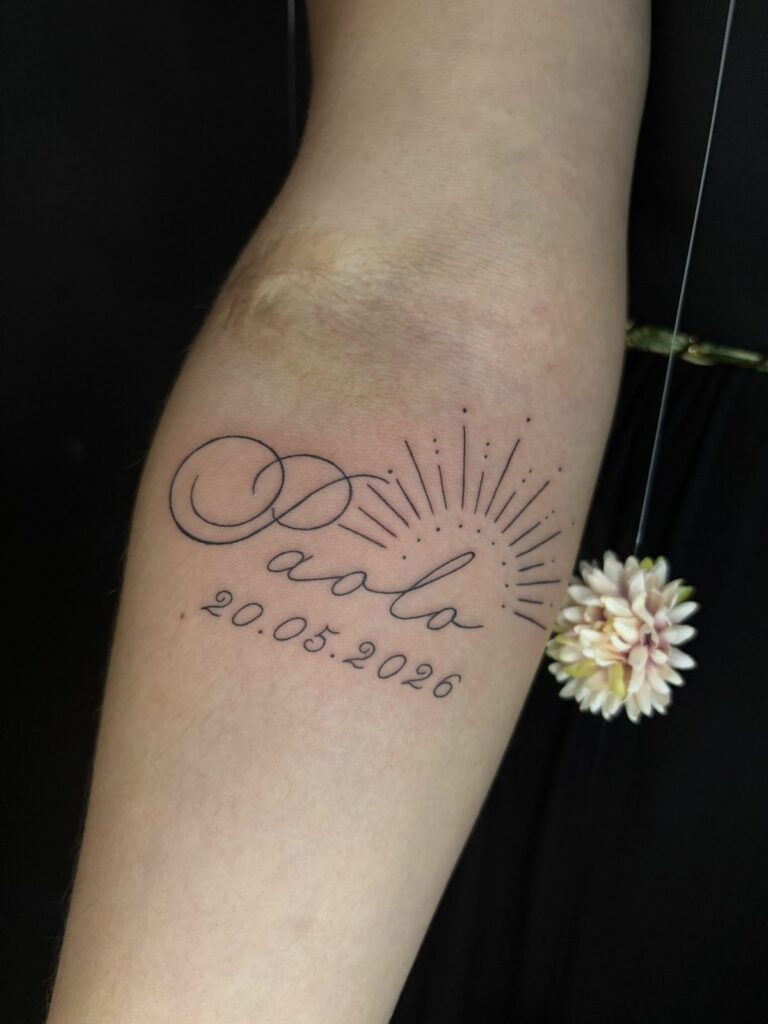 Tatouage fineline sur l’avant-bras : prénom en calligraphie à grandes boucles, date et rayons de soleil en traits et points, réalisé au salon Jolie Piqûre à Arles
