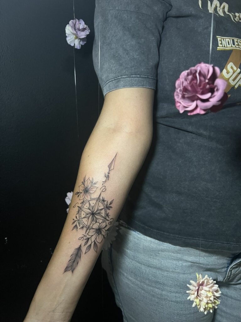 Tatouage fineline sur l’avant-bras : rose des vents entourée de fleurs et feuillage, avec flèche et plume, réalisé à Arles