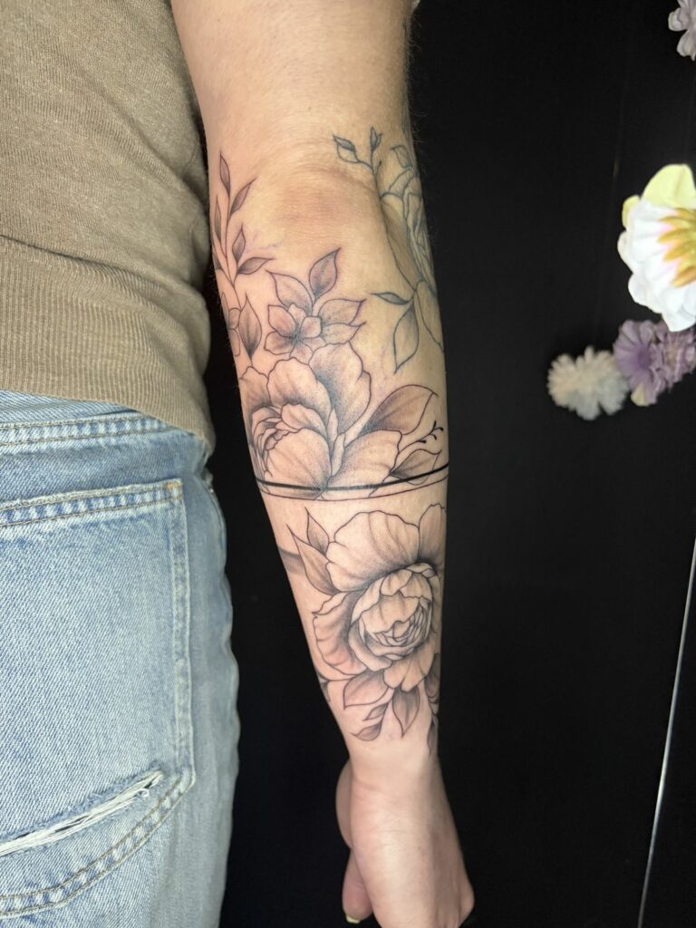 Tatouage fineline sur l’avant-bras : roses, pivoines et feuillages ombrés traversés par une ligne bracelet, réalisé au salon Jolie Piqûre à Arles