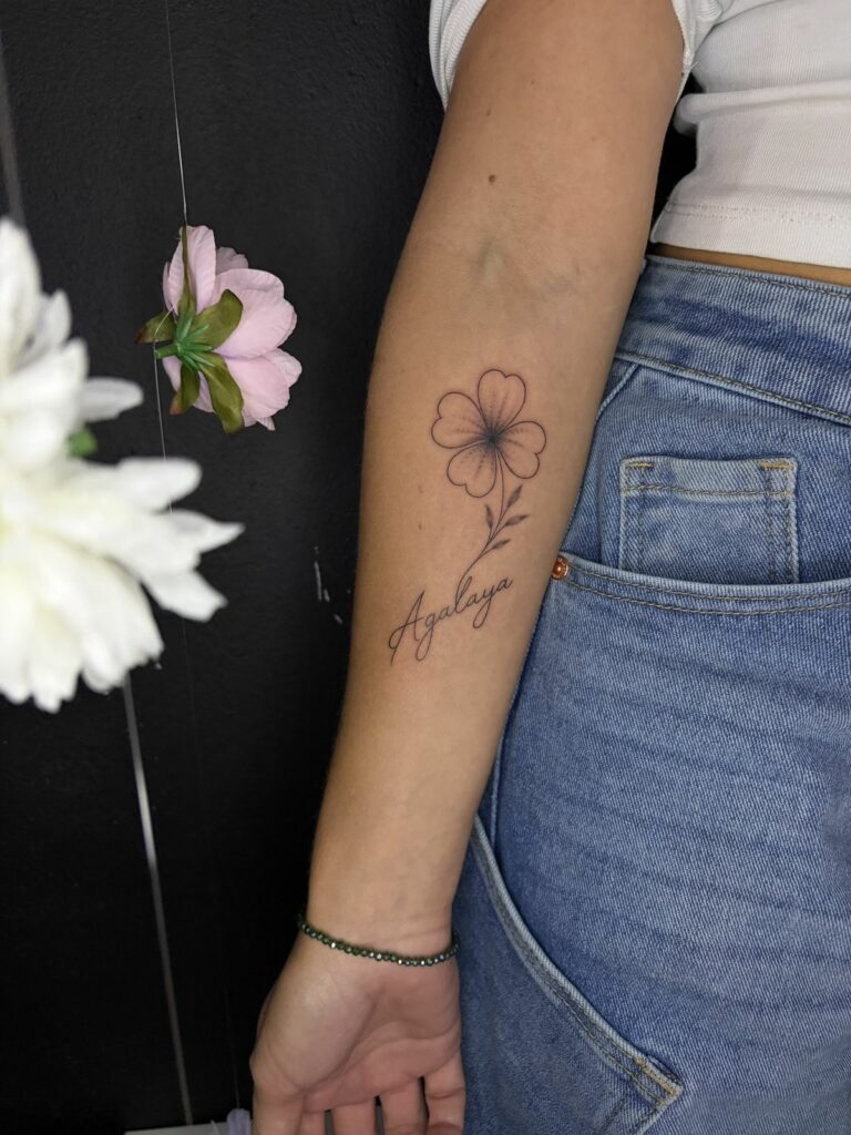 Tatouage fineline sur l’avant-bras : trèfle à quatre feuilles en pointillé avec tige feuillue et prénom en écriture manuscrite, réalisé au salon Jolie Piqûre à Arles