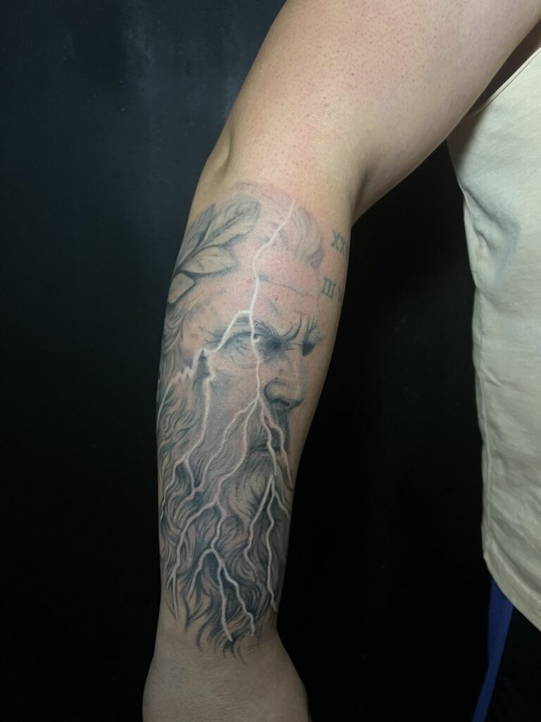 Tatouage réaliste noir et gris sur l’avant-bras : portrait de Zeus barbu couronné de laurier traversé d’éclairs blancs, réalisé à Arles
