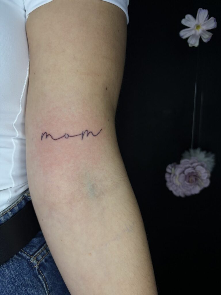Tatouage lettering fineline du mot « mom » en écriture manuscrite sur le bras, réalisé au salon Jolie Piqûre à Arles