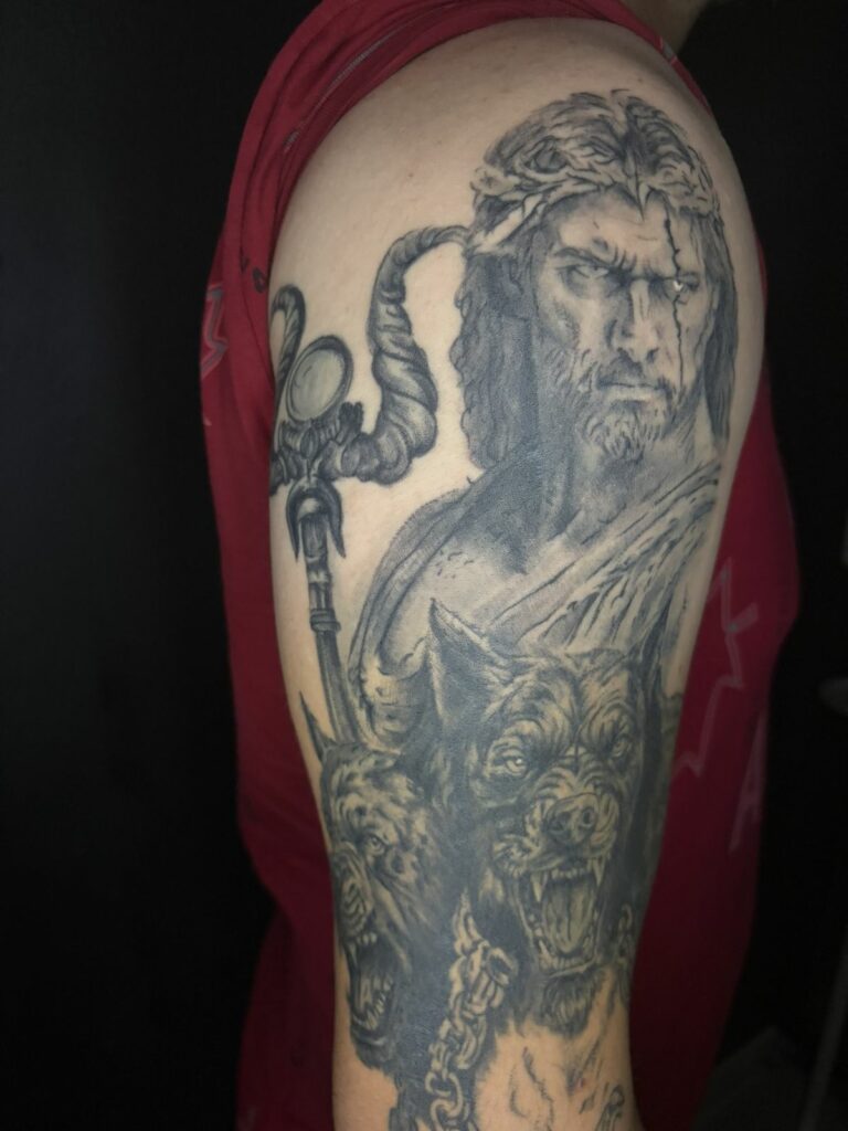 Tatouage réaliste noir et gris sur le bras : le dieu grec Hadès avec son sceptre et le chien à trois têtes Cerbère, réalisé au salon Jolie Piqûre à Arles