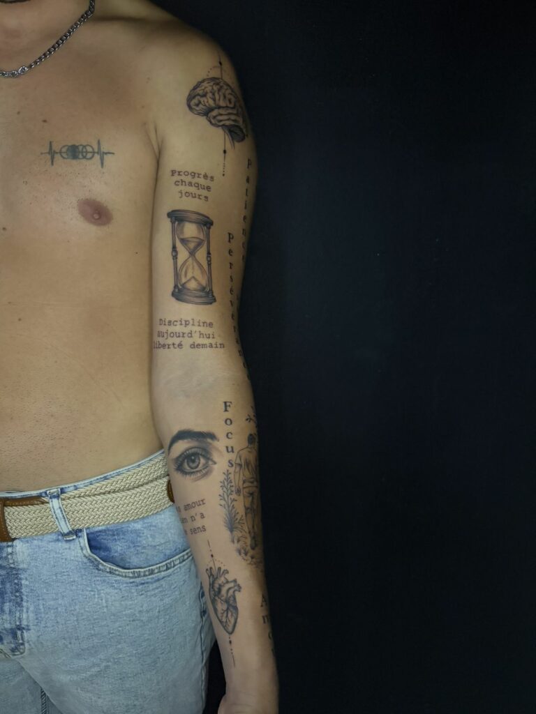 Tatouage patchwork noir et gris sur tout le bras : cerveau, sablier, œil réaliste, cœur anatomique et citations de motivation en lettrage machine à écrire, réalisé au salon Jolie Piqûre à Arles