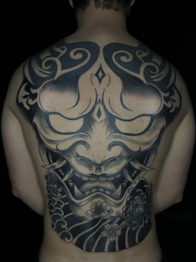 Grand tatouage de dos complet en noir et gris : masque Hannya de style japonais avec chrysanthème, fleurs de cerisier et vagues, réalisé au salon Jolie Piqûre à Arles