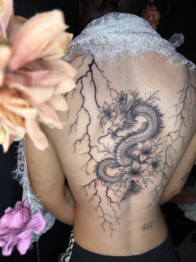 Grand tatouage sur le dos : dragon asiatique aux écailles détaillées entouré de fleurs de lys, avec des éclairs noirs sur les côtés, réalisé au salon Jolie Piqûre à Arles
