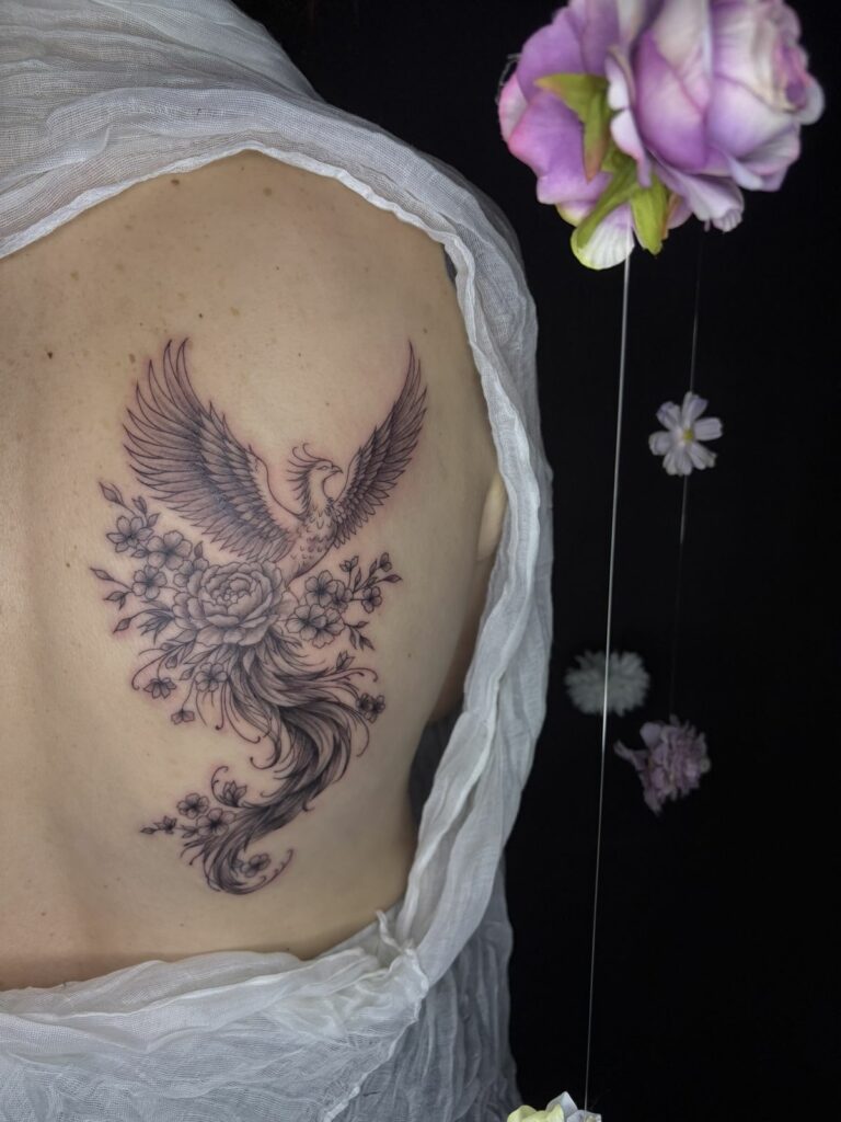 Tatouage fineline sur le dos : phénix aux ailes déployées entouré d’une pivoine et de fleurs de cerisier, réalisé au salon Jolie Piqûre à Arles