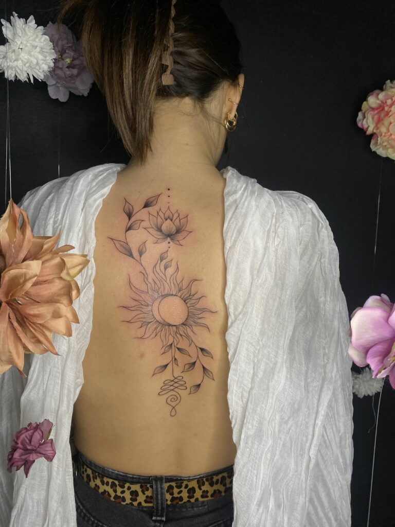 Tatouage fineline le long de la colonne : fleur de lotus, soleil et lune entrelacés, feuillage et symbole unalome, réalisé au salon Jolie Piqûre à Arles