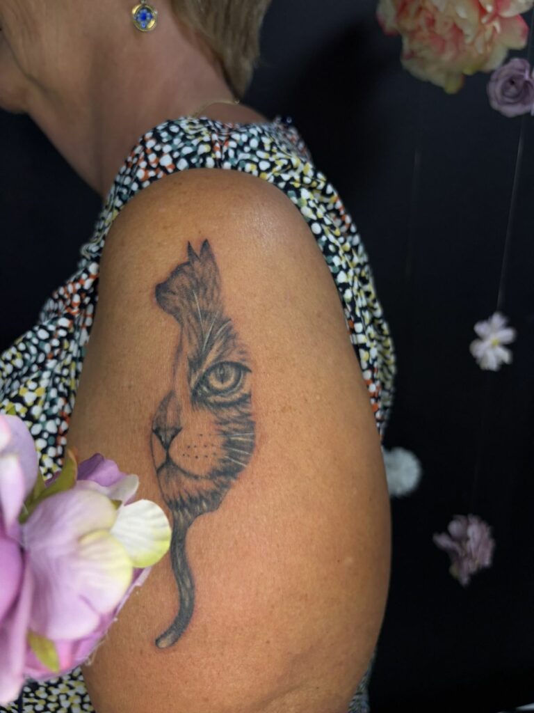 Tatouage réaliste noir et gris sur l’épaule : silhouette de chat assis intégrant le portrait détaillé d’un chat, réalisé au salon Jolie Piqûre à Arles