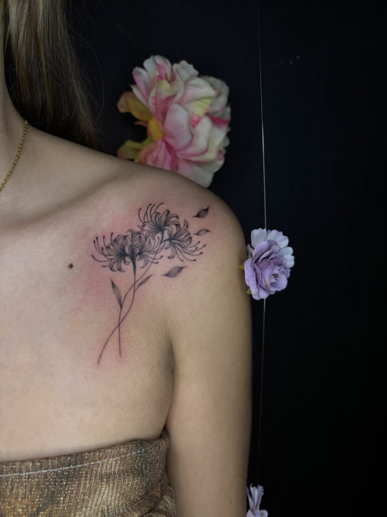 Tatouage fineline sur l’épaule et la clavicule : lys araignée aux longues étamines et pétales qui s’envolent, réalisé au salon Jolie Piqûre à Arles