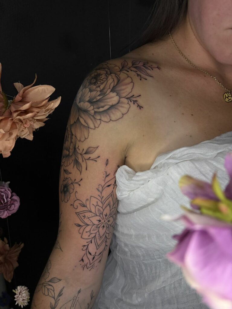 Tatouage fineline sur l’épaule et le bras : grande pivoine ombrée, petites fleurs et mandala ornemental en pointillés, réalisé au salon Jolie Piqûre à Arles