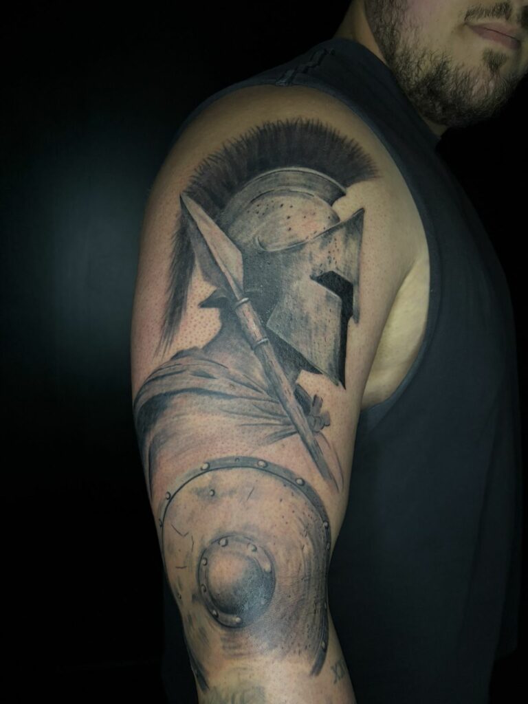 Tatouage réaliste noir et gris sur l’épaule et le bras : guerrier spartiate de profil avec casque à crête, lance et bouclier, réalisé au salon Jolie Piqûre à Arles