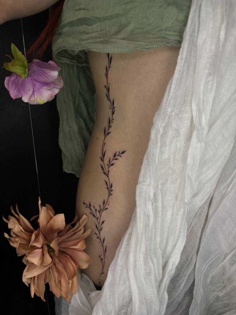 Tatouage fineline sur les côtes : longue liane de petites feuilles noires et points qui suit la courbe du corps, réalisé au salon Jolie Piqûre à Arles