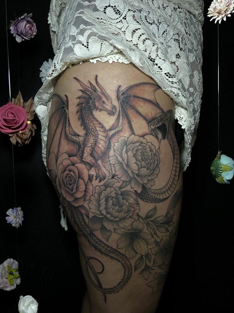 Grand tatouage noir et gris sur la hanche et la cuisse : dragon aux ailes déployées, serpent, roses et pivoines, réalisé à Arles