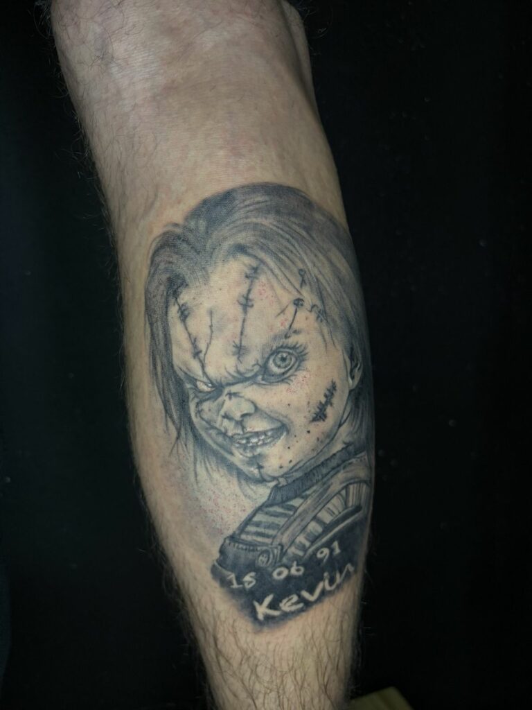 Tatouage réaliste noir et gris : portrait de la poupée Chucky du film d’horreur, avec cicatrices et sourire inquiétant, réalisé au salon Jolie Piqûre à Arles