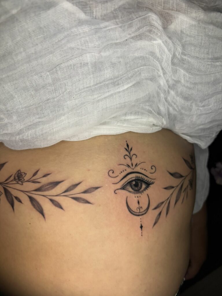 Tatouage ornemental sous la poitrine : œil réaliste, croissant de lune et ornements en pointillés, encadrés de branches feuillues, réalisé au salon Jolie Piqûre à Arles
