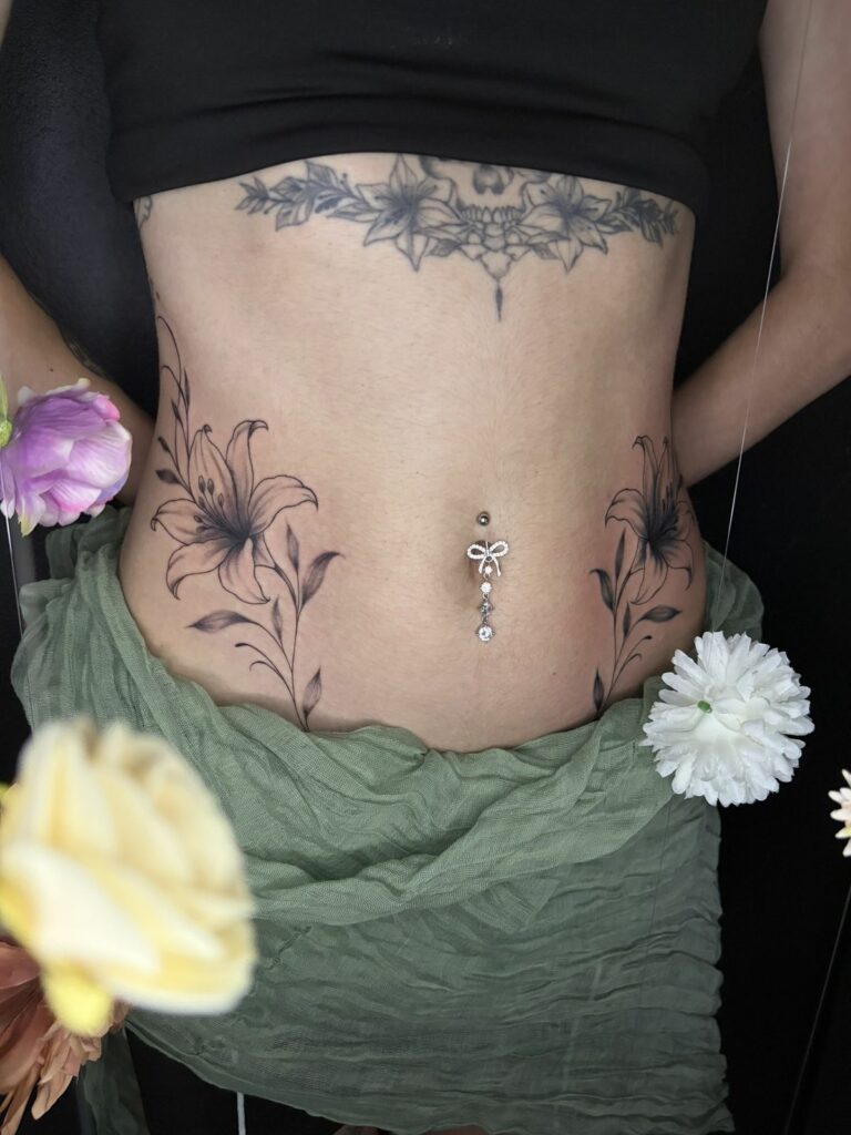 Tatouage fineline sur le ventre : deux fleurs de lys symétriques de chaque côté d’un piercing du nombril avec bijou nœud à strass, réalisé au salon Jolie Piqûre à Arles